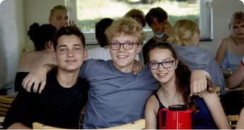 Tre teenagere sidder tæt sammen ved et bord og smiler til kameraet. De to drenge har armene om hinanden og pigen. Andre teenagere sidder i baggrunden og er i gang med en samtale.