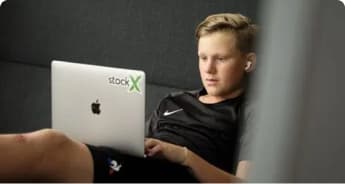 En ung person iført sort skjorte og trådløse øretelefoner sidder i en sofa og fokuserer på en sølvfarvet MacBook med et StockX-klistermærke på låget.