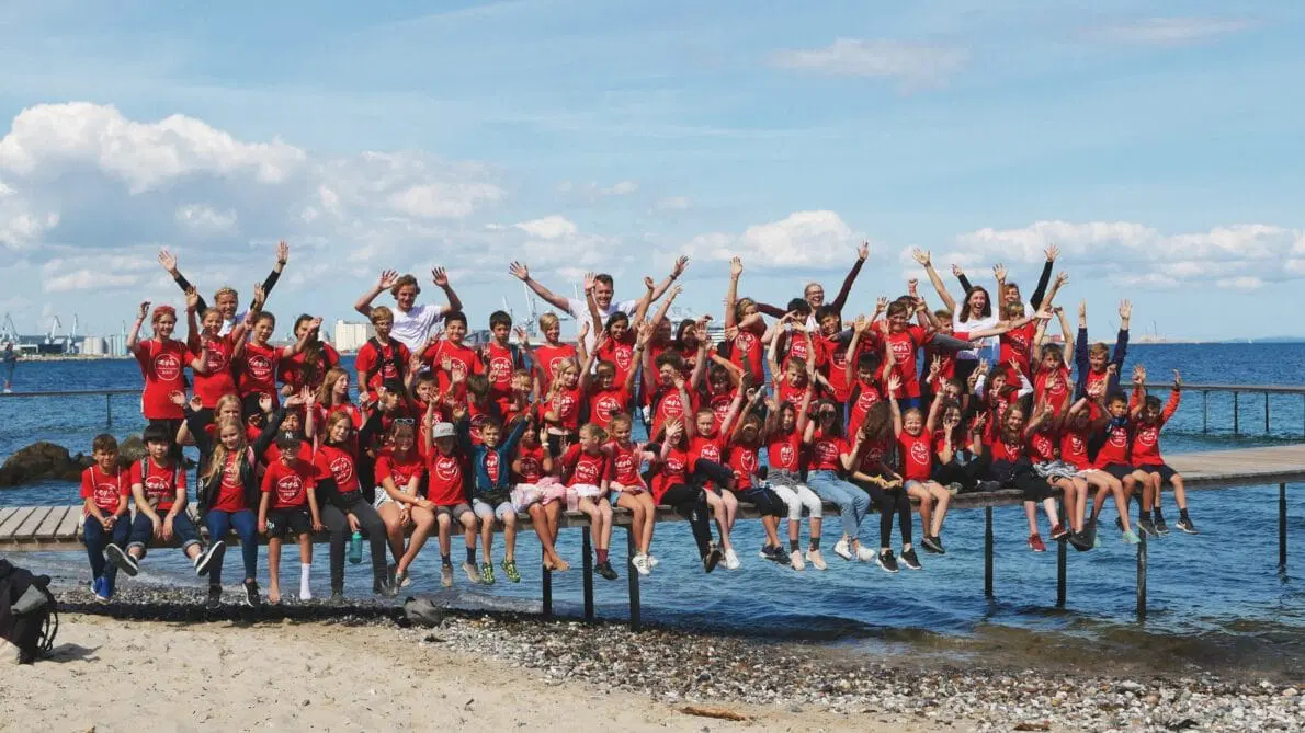 En stor gruppe børn og voksne i matchende røde skjorter sidder på en mole over vandet, smiler og løfter armene, med en strand, klipper og blå himmel i baggrunden.