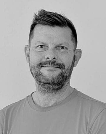 En mand med kort, stylet hår og skæg smiler let. Han er iført en almindelig t-shirt og er fotograferet i sort-hvid mod en enkel, lys baggrund.