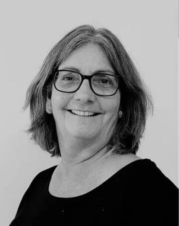 En kvinde med skulderlangt hår og briller smiler til kameraet. Hun er iført en mørk top, og billedet er i sort-hvid med en ensfarvet baggrund.