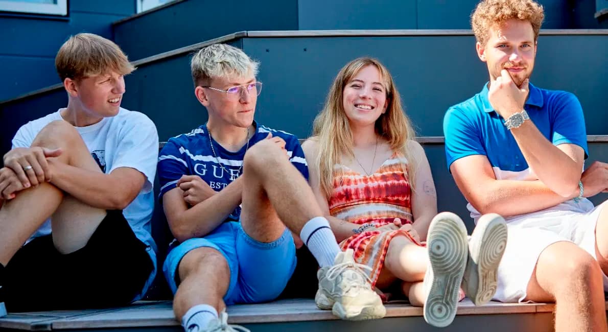 Fire unge voksne sidder på en udendørs trappe; tre mænd og en kvinde. Kvinden smiler og er iført et rødt batiktøj, mens de andre virker afslappede og er afslappet klædt. Det er en solrig dag.