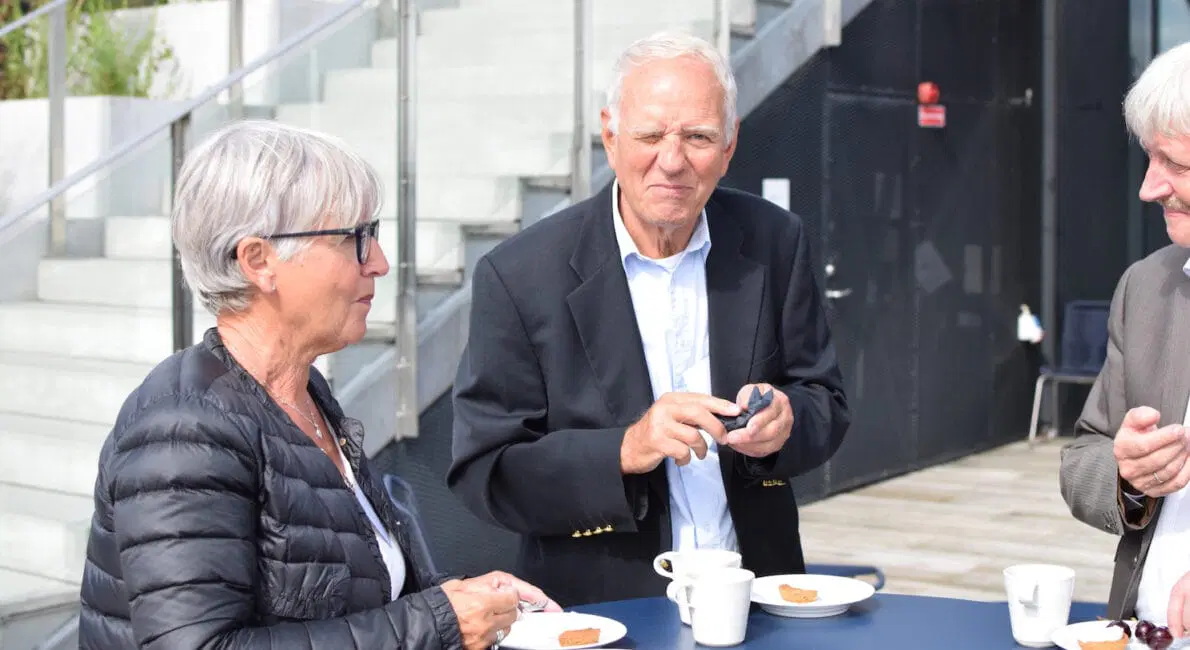 Tre ældre voksne står omkring et bord udendørs og drikker kaffe og spiser kager. To mænd har jakker på, en kvinde bærer briller og en sort jakke. Der er trapper og en indgang til en bygning i baggrunden.