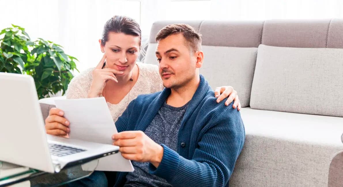 Et par sidder sammen indendørs og kigger på et dokument i mandens hånd, mens en bærbar computer er åben på bordet foran dem. Kvinden har sin hånd på mandens skulder, og begge virker fokuserede og eftertænksomme.