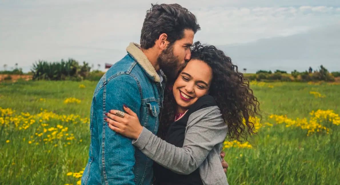 En mand kysser en smilende kvinde på panden, mens de omfavner hinanden på en mark med grønt græs og gule blomster under en overskyet himmel.