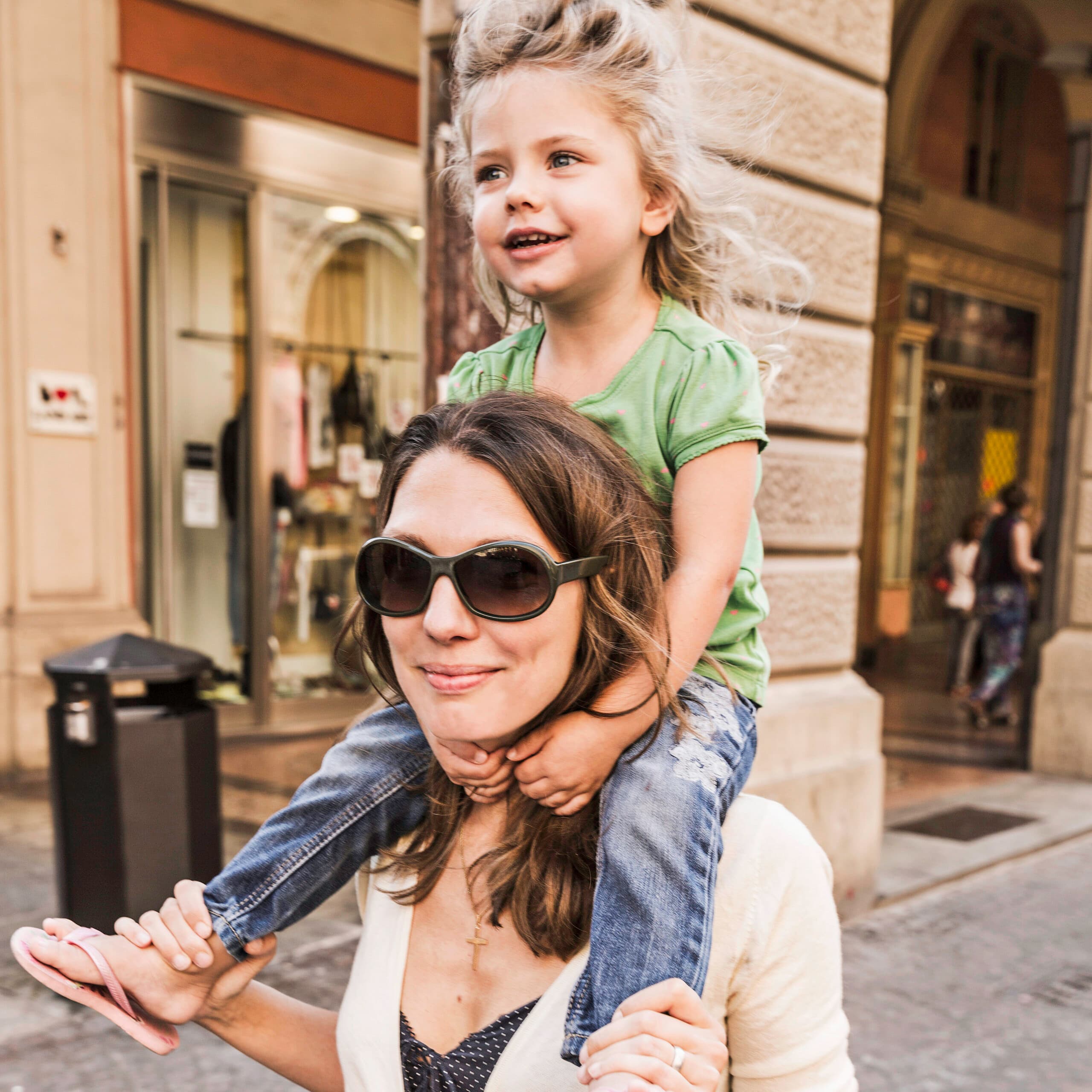 En kvinde med solbriller bærer en smilende ung pige på sine skuldre, mens de går ned ad en gade i byen med butikker og historiske bygninger.