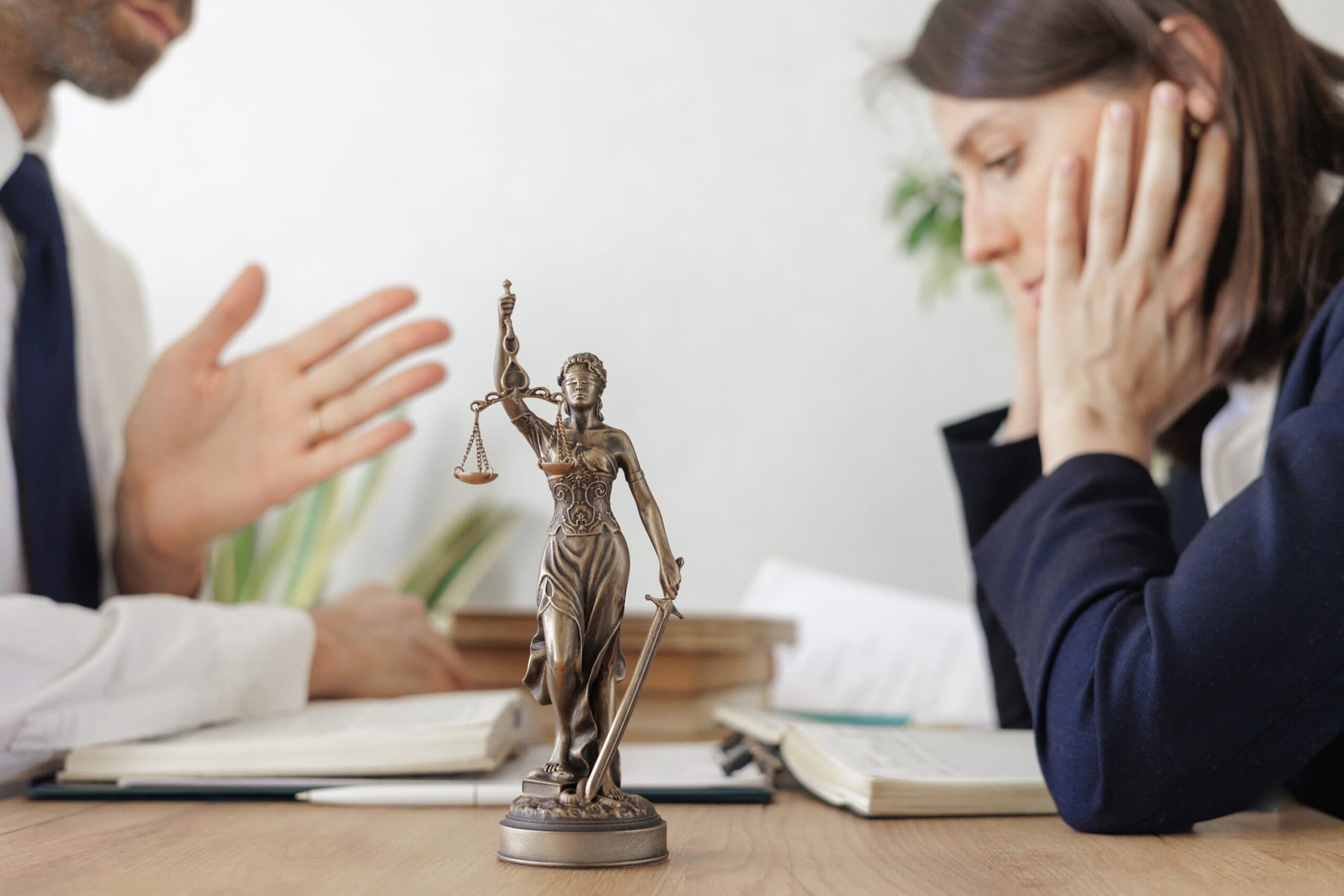 En statue af Lady Justice i messing sidder på et skrivebord mellem to personer i formelt tøj, som er i gang med en alvorlig samtale med åbne bøger og dokumenter foran sig.